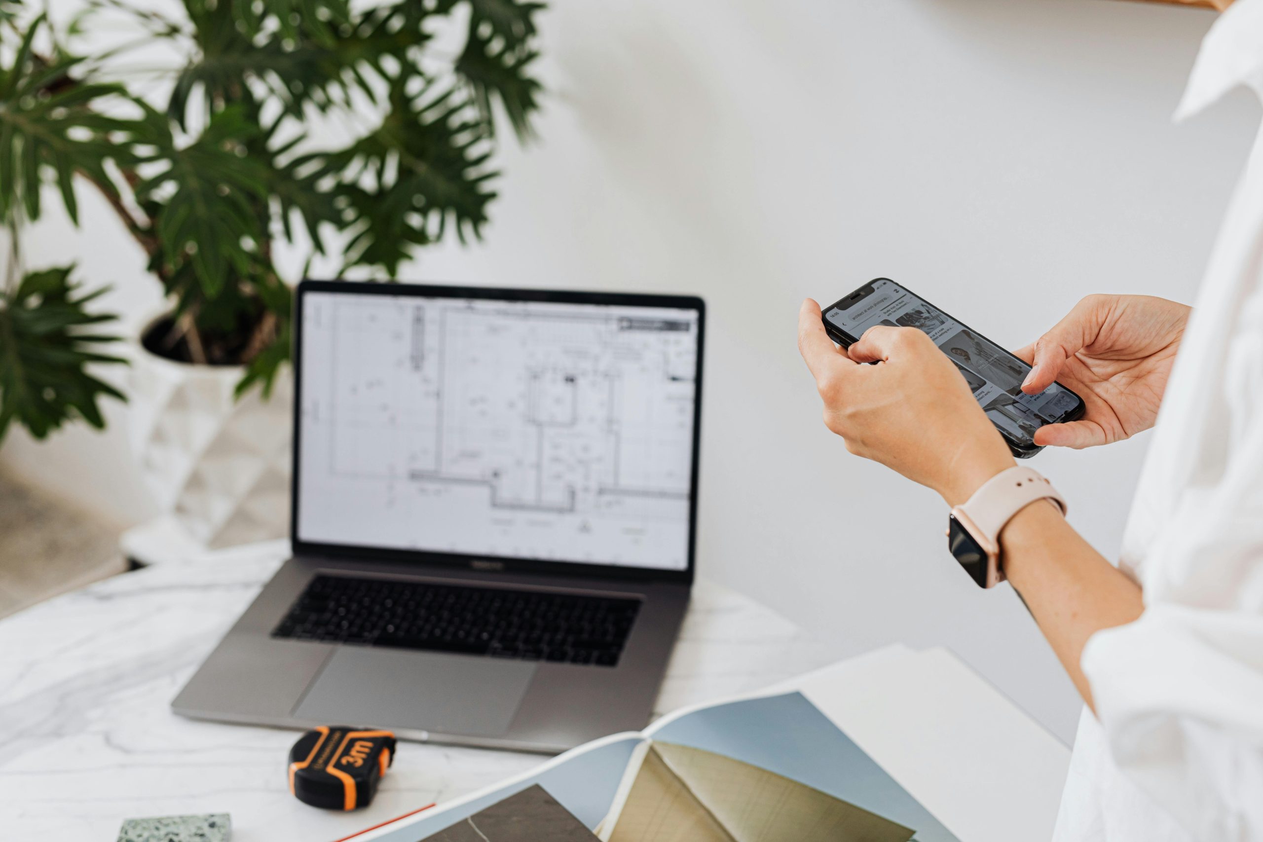 A person using a smartphone at a desk with a laptop displaying architectural plans.
